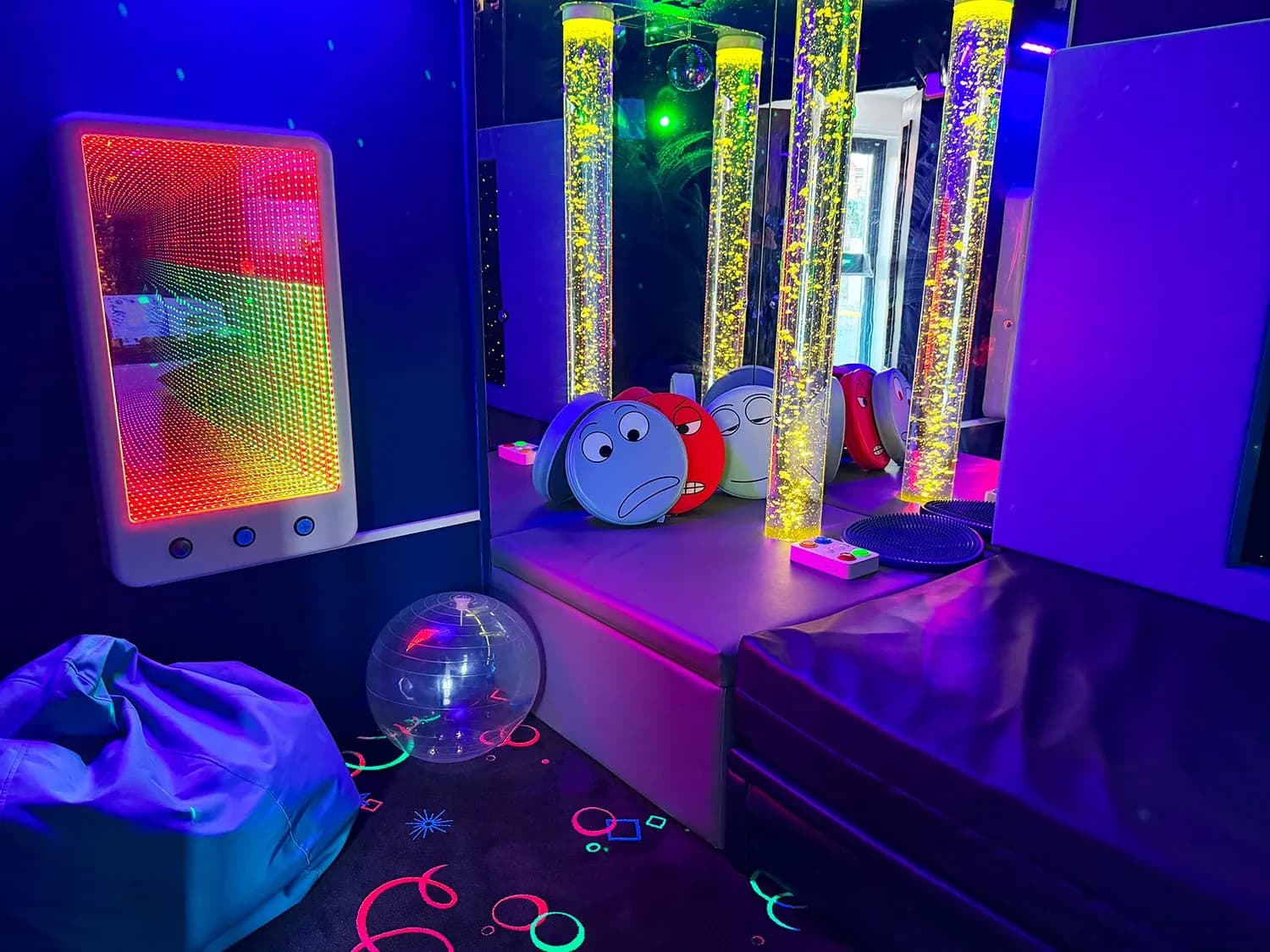 A sensory room features a glowing infinite mirror, bubble tubes, and cushions with cartoon faces, all bathed in blue and purple light with neon patterns on the floor.